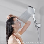 A person with long dark hair showers, holding a detached showerhead close to their head as water sprays down.