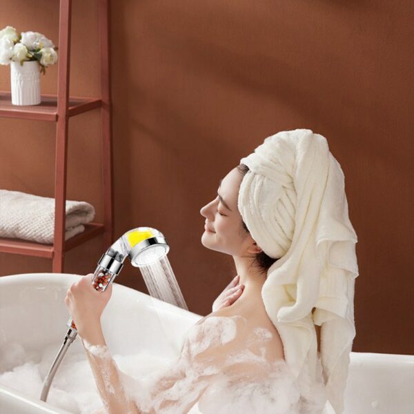 A person with a towel on their head enjoys a bubble bath, holding a showerhead in a brown-walled bathroom.