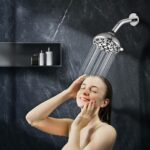 Person enjoying a shower under a stainless steel showerhead in a modern bathroom with dark marble walls and a toiletries shelf.