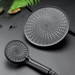 Black showerhead and handheld on a dark surface with a fern leaf and wet pebbles in the background.