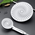 Two perforated showerheads, one larger, are displayed on a grey surface with a green fern and wet pebbles nearby.