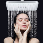 A person stands under a square showerhead, enjoying the water pouring onto their face and hands.