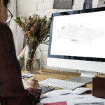 Someone sits at a desk with a computer showing a technical drawing, surrounded by papers, a keyboard, mouse, and a vase of dried flowers.