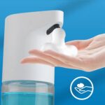 A hand under a white automatic soap dispenser releases foam. Blue background with a hand and foam icon in the corner.