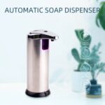 A stainless steel automatic soap dispenser with a black base and infrared sensor. Icons highlight features like induction, soap control, ABS material, and battery indicator.