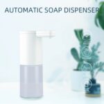 White automatic soap dispenser with icons for infrared sensor, non-contact use, and made of ABS. Background: blurred plants and soft lighting.