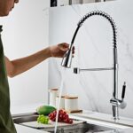 Person using flexible kitchen faucet to spray water over sink with grapes, mango, and white marble backsplash.