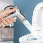 A hand aims a bidet spray at an open toilet bowl, water flowing.