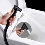 Hand holding a black bidet nozzle spraying water into a toilet, with a close-up inset of the nozzle.