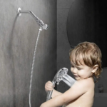 Un jeune enfant tient une pomme de douche dans la douche, avec un mur carrelé et un miroir rond derrière lui.