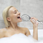 A person with short, wet hair enjoys a bubble bath, using a shower head as a microphone and singing.