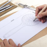 Person drawing a detailed showerhead illustration with pencil, drafting tools on the table.