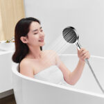 A person in a bathtub directs the showerhead spray onto their face.