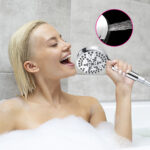 A short-haired blonde person in a bubble bath holds a showerhead near their mouth, with an inset showing water spraying from it.