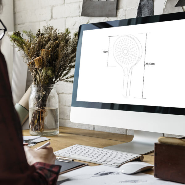 Personne au bureau regardant le design de la douchette à main avec des mesures sur ordinateur, entourée de fournitures de bureau et d'un vase de fleurs séchées.