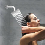 A wet-haired person stands under a shower, hands behind their head, water flowing from a square showerhead on the tiled wall.