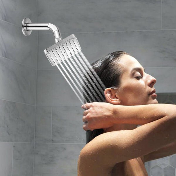 A wet-haired person stands under a shower, hands behind their head, water flowing from a square showerhead on the tiled wall.