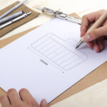 Person drawing a technical design on paper with a pencil, surrounded by drawing tools like a ruler, eraser, and mechanical pencil.