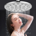 A woman stands under a large showerhead, water streaming down. She smiles slightly with her eyes closed, enjoying the shower.