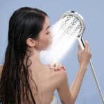 A woman with wet hair rinses her shoulder using a filtered handheld shower head against a light blue background.