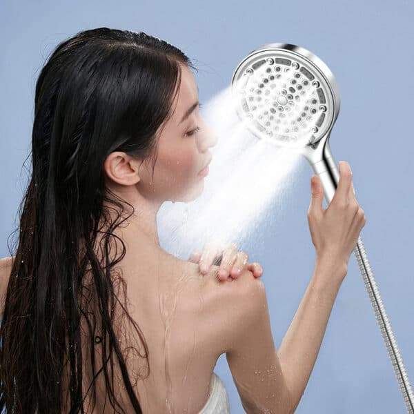 A woman with wet hair rinses her shoulder using a filtered handheld shower head against a light blue background.