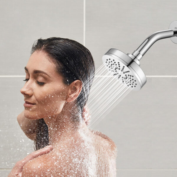 Una mujer con el cabello mojado se enjuaga la espalda y el cuello bajo una gran ducha redonda montada en la pared en un baño de azulejos.
