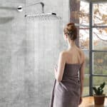 Une femme en chignon, enveloppée dans une serviette, se tient dans une salle de bain carrelée face à une grande douche à effet pluie Leelongs près de grandes fenêtres.