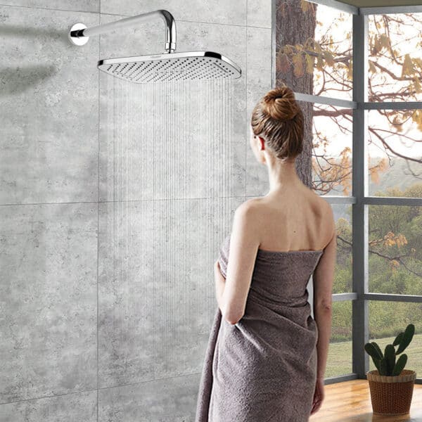 Une femme en chignon, enveloppée dans une serviette, se tient dans une salle de bain carrelée face à une grande douche à effet pluie Leelongs près de grandes fenêtres.