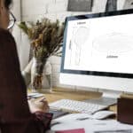 Persona en un escritorio con una computadora que muestra diagramas del cabezal de ducha de 9 pulgadas de Leelongs, útiles de oficina y flores secas cerca.