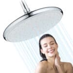 壁に取り付けられた丸いレインシャワーヘッドの下で、女性が目を閉じて微笑んでいます。シャワーヘッドから水が降り注いでいます。