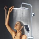 Une femme se tient sous une grande douche à effet pluie chromée, les yeux fermés, le bras levé ; une douche à main est également fixée.