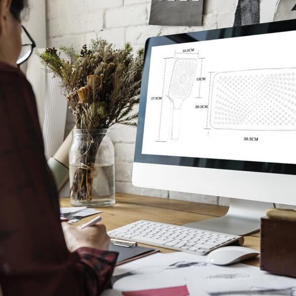 Uma pessoa sentada em uma mesa visualiza desenhos técnicos e medidas de um chuveiro na tela do computador.