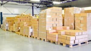 Des boîtes en carton emballées dans du plastique sont posées sur des palettes dans un entrepôt avec un sol en béton et un haut plafond.
