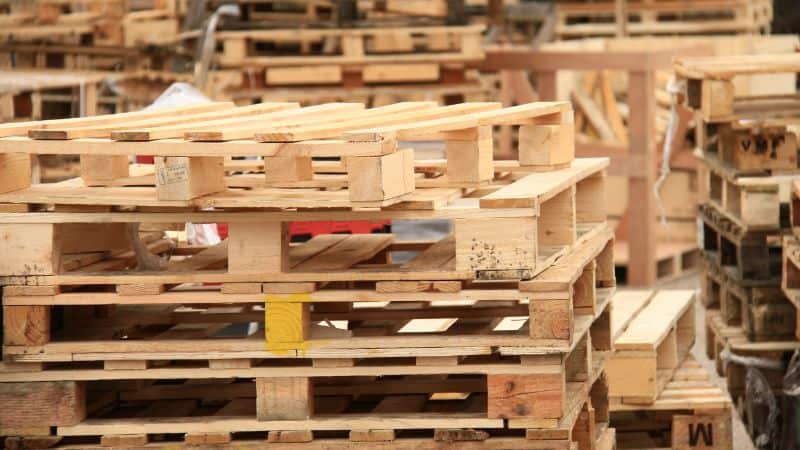 Des piles de palettes en bois de différentes tailles et couleurs sont stockées à l'extérieur.