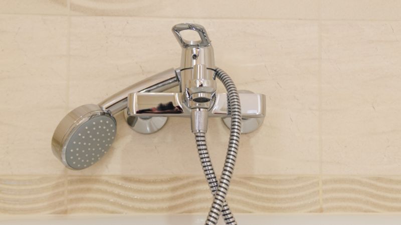Shower faucet with handheld spray hose mounted on a tiled bathroom wall, modern chrome finish, designed for versatile water use and easy cleaning.