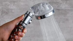 Alt: Hand holding a modern shower head with water flowing, featuring colorful bath beads inside the handle, against a gray textured background, showcasing innovative bathroom accessories.