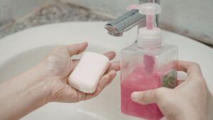 El hijyenini ve sabun türlerini gösteren, lavabo yakınında pompalı sabunluktan köpük el sabunu ve kalıp sabun kullanan ellerin yakın çekim görüntüsü.