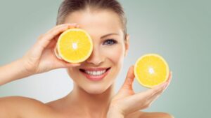 شابة تحمل نصفي ليمون طازجين على عينيها، تبتسم ابتسامة مشرقة، تروج لدور فيتامين سي في صحة الجلد والشعر.