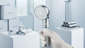 Un moderno soffione doccia cromato, tenuto da una mano guantata, si trova in un bagno pulito, con sanitari e asciugamani arrotolati sullo sfondo.
