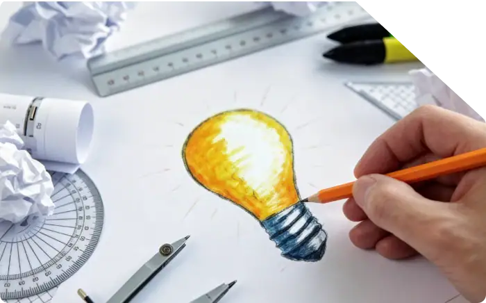 Hand drawing a colorful lightbulb sketch on paper, symbolizing ideas and creativity, with drafting tools and crumpled paper around.