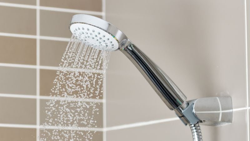 Gros plan d’un pommeau de douche moderne en fonctionnement dans une salle de bains aux murs carrelés beiges.