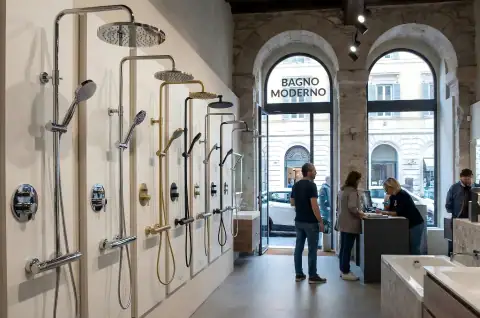 Showroom interior featuring a variety of modern showerheads and fixtures displayed on a wall, with customers browsing and a large window in the background.
