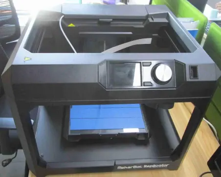 3D printer on a black stand with a transparent cover, showing the printing area and control panel, used for creating prototypes and models.